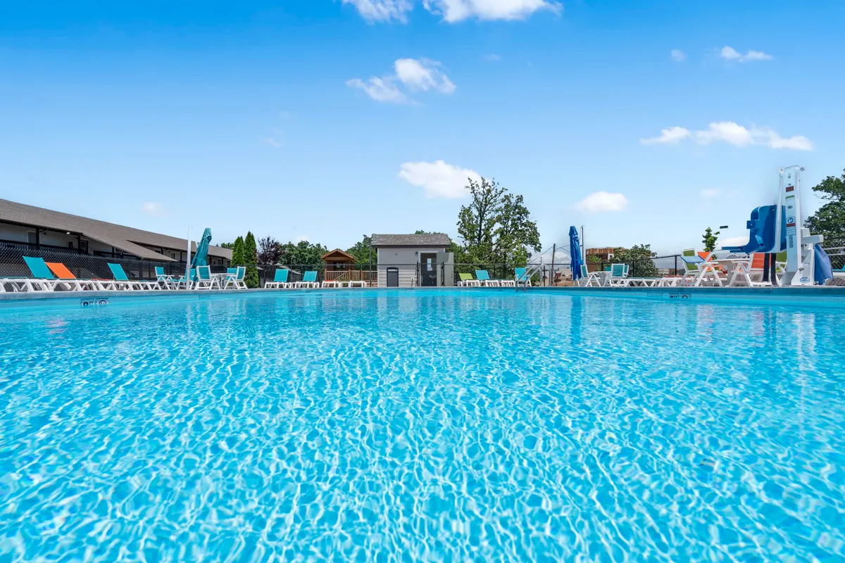 This lively outdoor pool scene features a large swimming pool surrounded by colorful lounge chairs, a waterslide, and a lifeguard station. People are enjoying sunny afternoons swimming, socializing, and making memories.