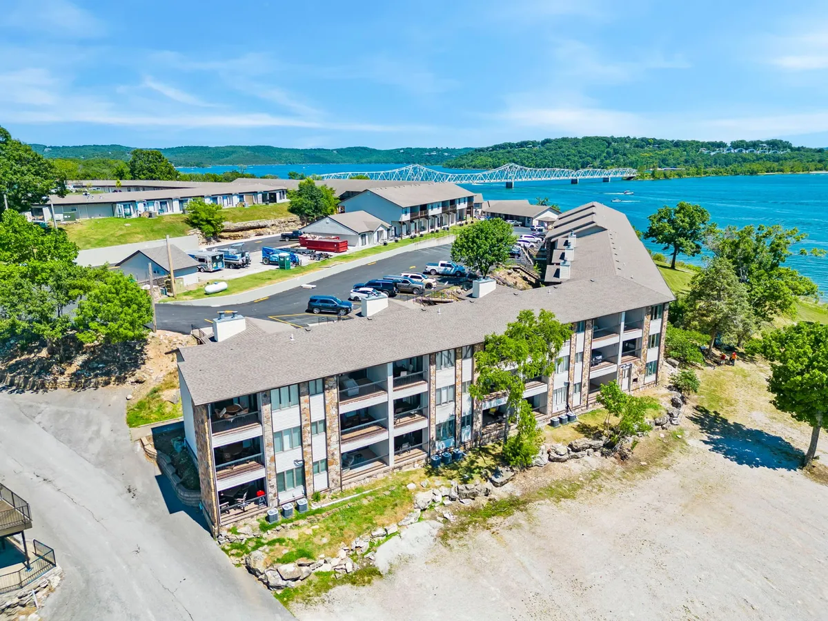 This image captures a multi-story building set against a lush backdrop of green trees, with Table Rock Lake visible in the scene. The mood is relaxed, and the setting sun casts a warm glow over the water. People can be seen boating, fishing, or picnicking along the lake's edge, perfectly representing the lakeside lifestyle and the activities available just steps from the property.