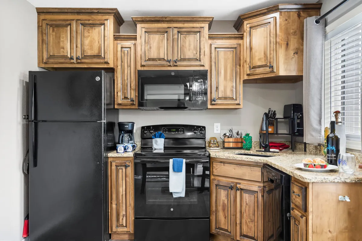 A cozy and functional kitchen featuring wooden cabinets, sleek black appliances, granite countertops, and a dedicated coffee station. The window with curtains allows for natural light, brightening up the space. The layout provides ample counter space for meal prep and group cooking, while the modern appliances make entertaining and daily meal routines effortless.