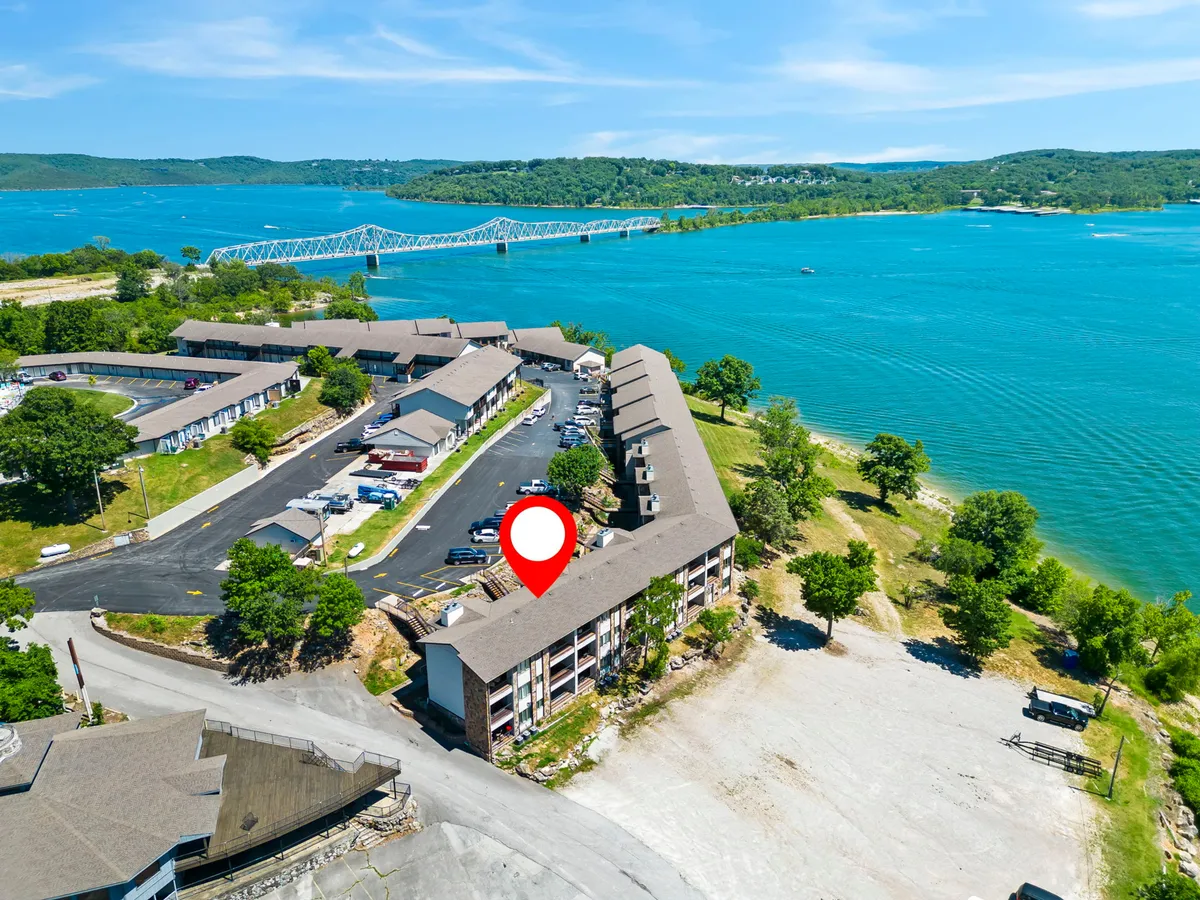 A sweeping waterfront view showcases a calm lake bordered by lush greenery and nearby buildings. The scene captures the tranquility of the setting, with space for boating, fishing, and relaxing moments by the water. The parking area and buildings in the distance indicate accessibility and a welcoming environment for guests year-round.