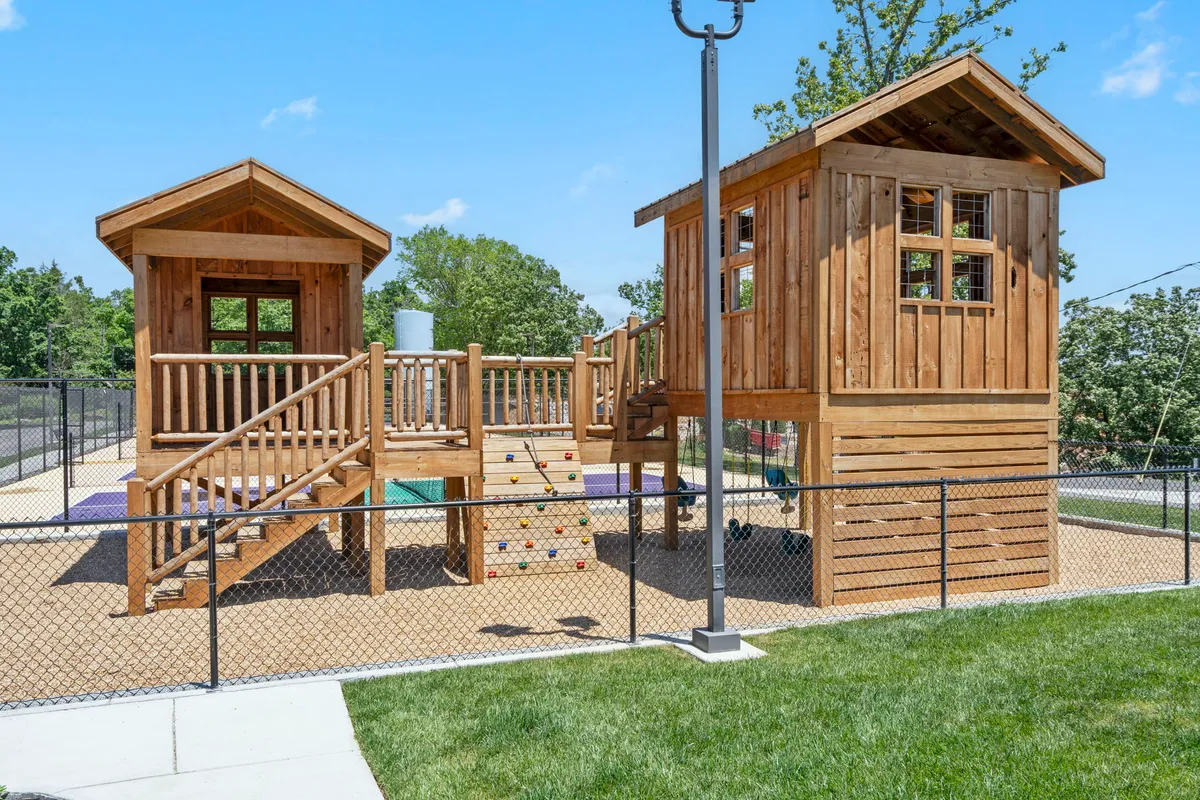 A lively playground scene with a playhouse structure, climbing wall, slide, and swings. Children can be seen playing and exploring, with plenty of space for imaginative outdoor adventures.