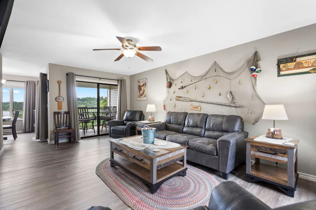 The image showcases a warm, inviting living area in the condo, featuring plush leather sofas arranged for socializing, a coffee table perfect for board games, and a decorative fishing net that nods to the lakeside setting. Through the windows, the soft glow of a sunset hints at balcony views of Table Rock Lake.