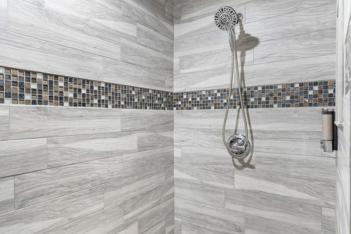 A stylish bathroom with a modern showerhead, decorative tile strip, and wood-like wall tiles. The clean and serene design highlights the spa-like feeling of the ensuite bath.