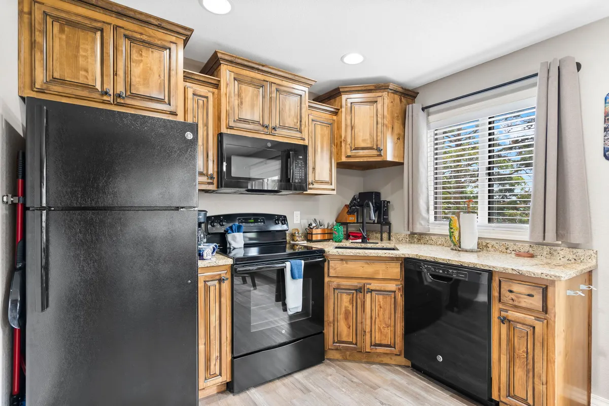 A second perspective of the kitchen, emphasizing the wooden cabinetry, black appliances, and granite countertops. Natural light streams through the window, creating a welcoming environment suited for both casual breakfasts and elaborate meal preparations.